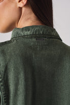 Close-up of the branded metal tag on the collar of the green denim shirt.