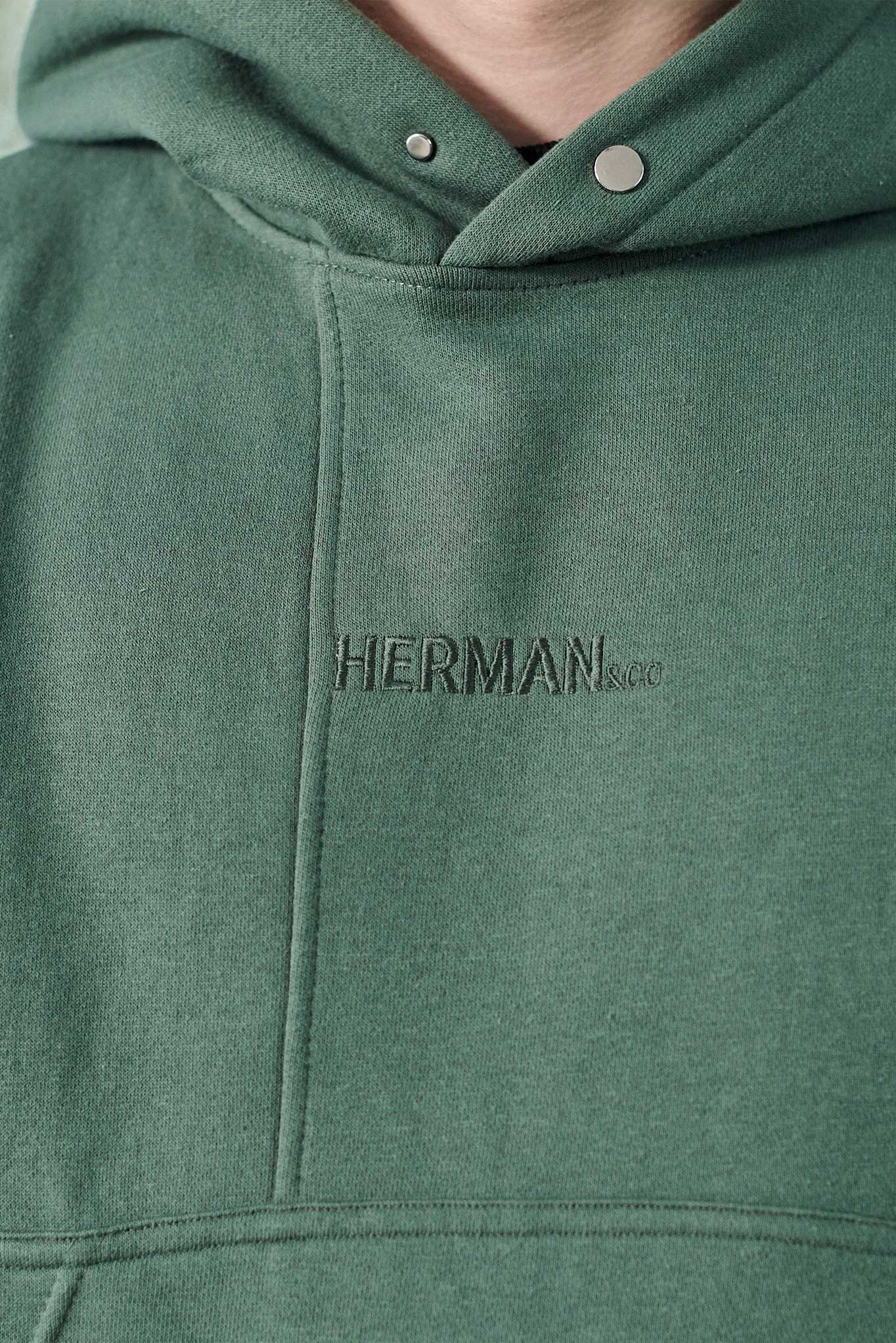 Detail of the tonal HERMAN logo embroidery on dark green hoodie chest